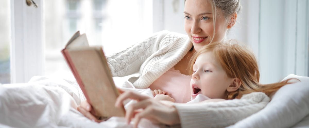 Cheerful young woman hugging cute little girl and reading book together while lying in soft bed in light bedroom at home in daytime